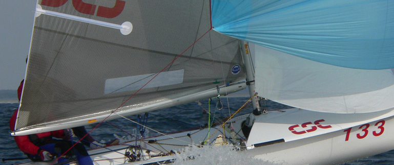 Close-up of a white sailboat with the number 133 on the hull, cutting through the water with its sails fully open and a person in a red jacket visible behind the sail.