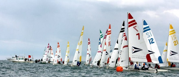 A row of small sailboats with colorful, numbered sails line up at the start of a race on the water, with a boat nearby and cloudy sky overhead.