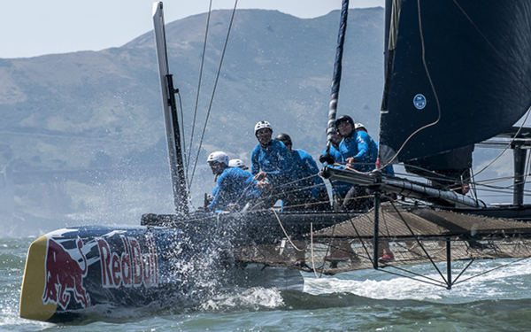 A team of sailors wearing blue gear race on a Red Bull-branded catamaran, cutting through choppy water with sprays splashing up, against a backdrop of hills and a partly cloudy sky.
