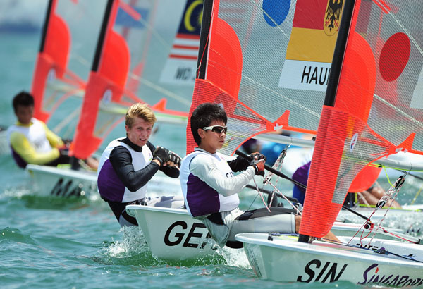 Four sailors compete in a sailing race, each in their own small sailboat with colorful sails featuring country flags. They wear life vests and sunglasses as they steer their boats across the water.