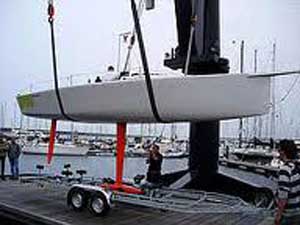 A white sailboat is being lifted by a crane onto a trailer at a marina, with several people nearby and other boats docked in the background.