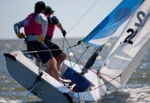 Two sailors wearing life jackets maneuver a small sailboat, leaning out over the water as the boat tilts. The blue-and-white sail is up, and the water is choppy under a clear sky.