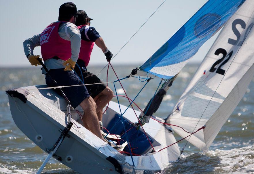 Two sailors wearing life jackets maneuver a small sailboat, leaning out over the water as the boat tilts. The blue-and-white sail is up, and the water is choppy under a clear sky.