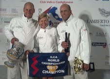 Three people in white jackets stand together, smiling and holding trophies and a banner that reads 'ILCA World Championship 2013 Champion,' with a backdrop of sponsor logos behind them.
