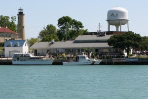 Two boats are docked along a waterfront with a lighthouse, trees, a large water tower, and several buildings in the background under a clear sky.