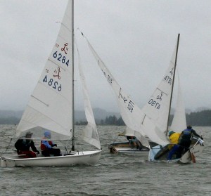 Four small sailboats are racing on a gray, choppy lake under overcast skies. Two boats appear to be capsizing, while sailors in rain gear try to stabilize them. The scene looks cold and windy.