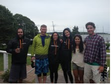 Five people stand outdoors, smiling at the camera. Four wear casual jackets or hoodies, while one wears a plaid shirt. Trees and a building are visible in the background.