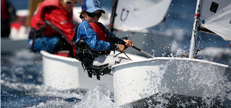 A young sailor wearing a blue shirt, life jacket, and cap steers a small sailboat through splashing water, with another sailboat and sailor visible in the background on a sunny day.