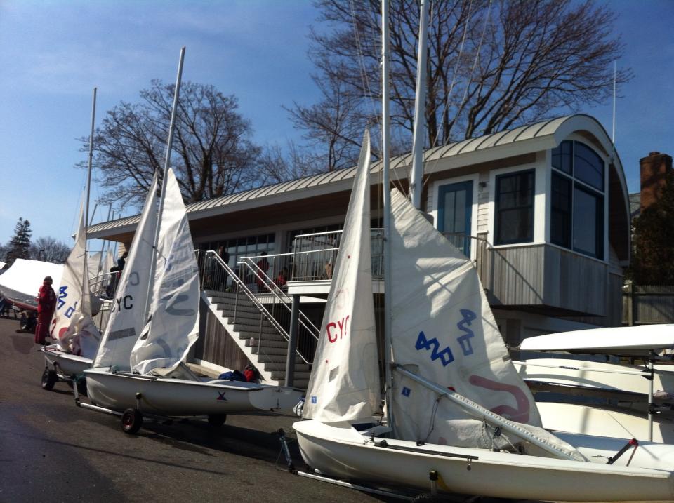 Three small sailboats with white sails are parked on land in front of a light gray boathouse with large windows and stairs. Leafless trees and clear blue sky are in the background.