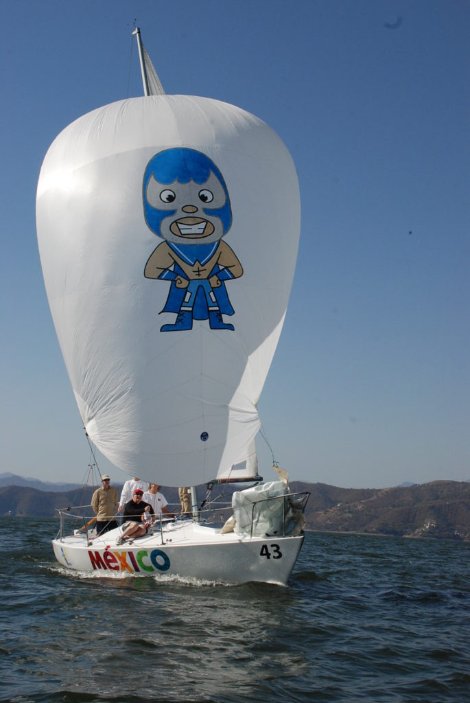 A white sailboat with 'Mexico' painted on the side sails on a lake. The large sail features a cartoon luchador in a blue mask. Several people are onboard, and mountains are visible in the background.