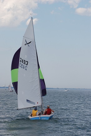 Two people sail a small boat with a white sail marked “2493” on a calm body of water under a partly cloudy sky. The boat has a green and purple spinnaker sail. Other boats are visible in the distance.