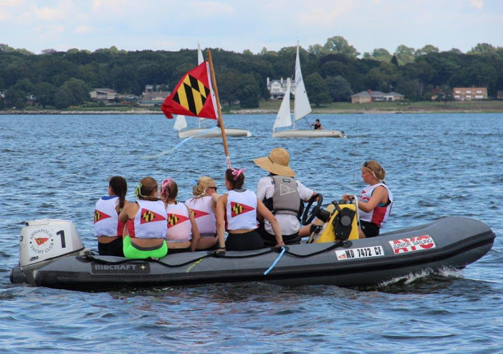 A group of people in matching vests ride in a small inflatable boat on a lake, holding a Maryland flag. Sailboats and a tree-lined shore with houses are visible in the background.