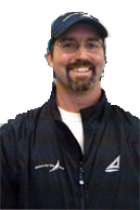 A smiling man with glasses, a beard, and a mustache wears a navy baseball cap and a navy jacket with a triangular logo on the left side.