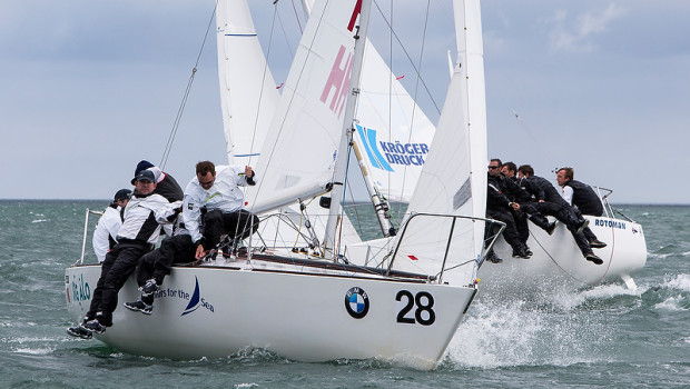 Two racing sailboats with crews in black and white gear lean out over the side, competing closely in choppy waters under a cloudy sky. The boat in front is numbered 28 and has visible sponsor logos on its hull and sails.