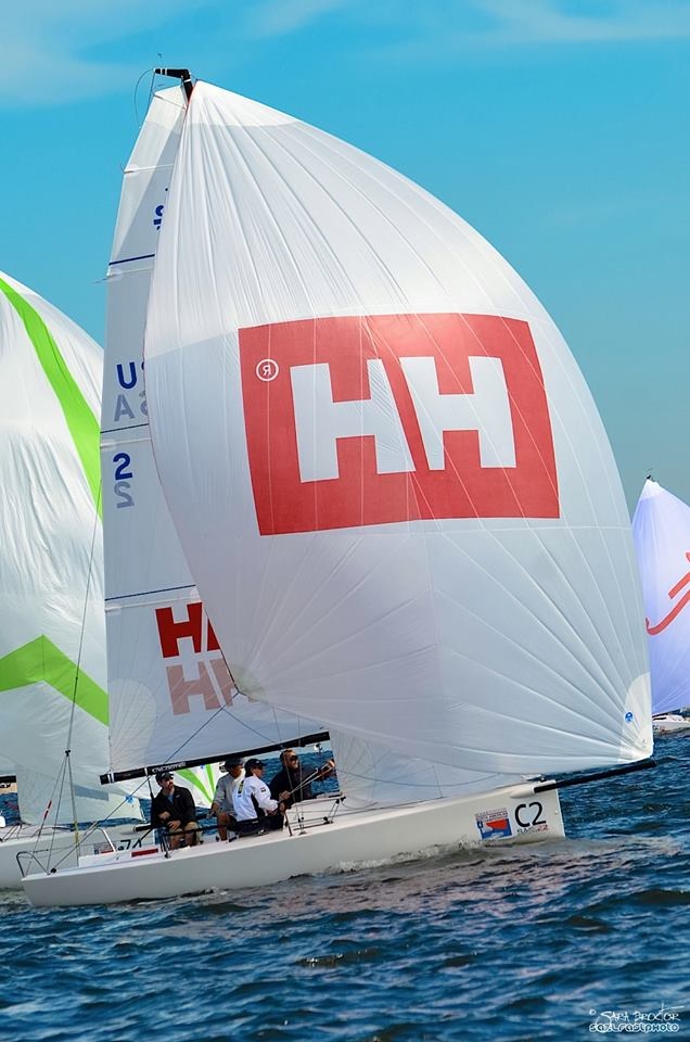 A white sailboat with a large Helly Hansen (HH) logo on its sail glides over blue water. Several people are on board, and other sailboats are visible in the background under a bright sky.