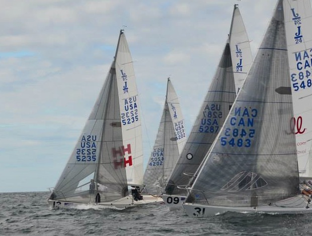 Several sailboats with white sails, marked with 'USA' and 'CAN' and various numbers, race closely together on choppy water under a cloudy sky.