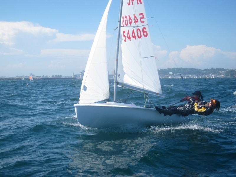 Two people sail a small white sailboat with the sail marked 'JPN 54406' on choppy blue water, leaning out over the edge. The background shows coastline and partly cloudy skies.