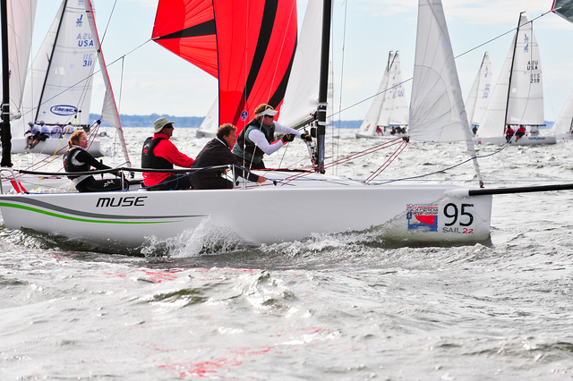 Four people are sailing a white boat labeled 'MUSE' and '95' in a regatta, with other sailboats visible in the background on a choppy body of water. The sails are red, black, and white.