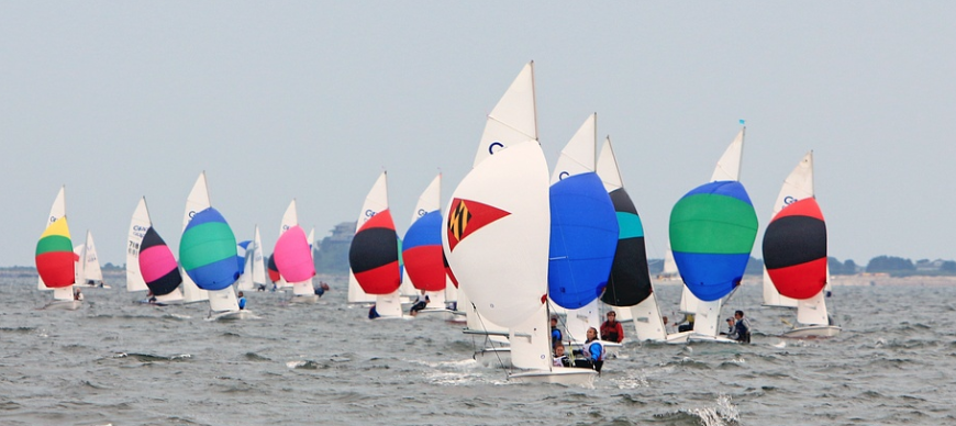 A group of colorful sailboats with spinnakers open races on a slightly wavy body of water, with distant land and buildings visible on the horizon under a cloudy sky.