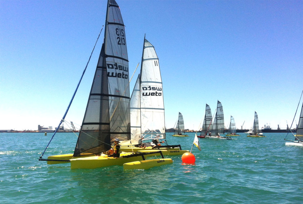 Several small yellow trimaran sailboats race on a bright blue sea, passing a red buoy under a clear blue sky. Other sailboats are visible in the background, along with distant ships and a coastline.