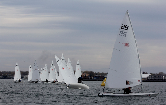 Several small sailboats with white sails race on a gray, overcast day. One boat in the center is capsized, its hull visible above the water. Other boats and shoreline buildings are in the background.