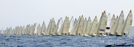 A large fleet of sailboats with white sails lined up closely together on the open water, participating in a sailing race under a hazy sky.