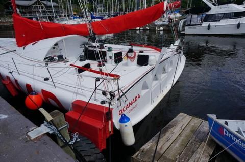 A white sailboat with red accents named 'O CANADA' is docked at a marina, surrounded by other boats. The boat has a red sail and various equipment on deck. A blue dinghy is tied to the dock nearby.