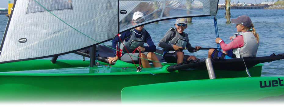 Three people wearing life jackets and sunglasses sit in a bright green sailboat on the water, smiling and talking, with sails up and a shoreline visible in the background.