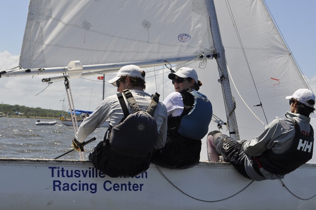 Three people wearing life jackets and caps are sitting on the edge of a sailboat marked 'Titusville Yacht Racing Center,' sailing on a body of water under a partly cloudy sky.