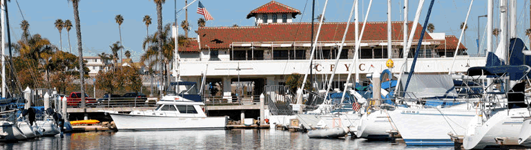 Boats and sailboats are docked at a marina in front of a building with a red-tiled roof, palm trees, and an American flag. The building's sign reads 'CYC.