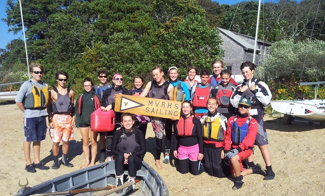 A group of teens wearing life jackets stands on a sandy shore, smiling and holding a wooden “MVRHS Sailing” sign. Sailboats and trees are in the background, with a house partially visible behind them.