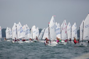 A group of young sailors in small white sailboats compete in a regatta on a calm body of water under a cloudy sky, with many sails visible and participants wearing life jackets.