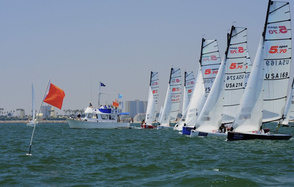 Several sailboats with white sails line up at the start of a race near a motorboat, with a red flag on a buoy in the foreground and city buildings visible in the background on a clear day.