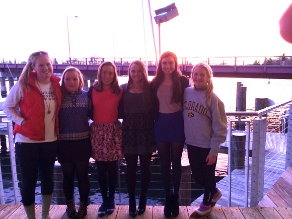 Six young women stand side by side on a wooden deck by the water at sunset, with a bridge and railing in the background. They are smiling and dressed in casual to semi-formal attire.