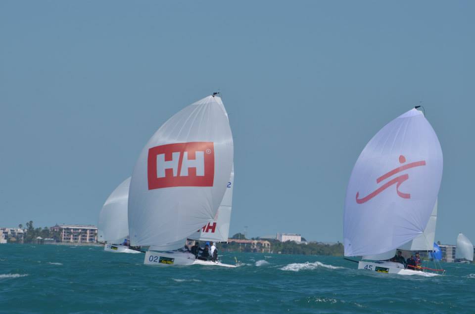 Two sailboats with large white sails race on the ocean; one sail features a red 'HH' logo, and the other has a stylized figure in red. Buildings and trees are visible on the distant shoreline under a clear sky.