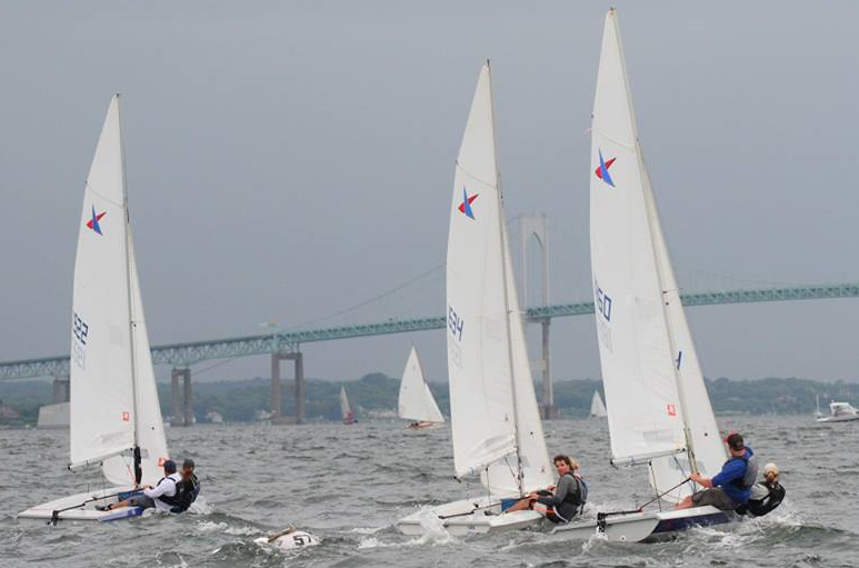 Several small sailboats with people aboard race on choppy water, with a large bridge and more boats in the background under a cloudy sky.