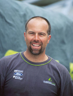 A man with a beard and short hair smiles at the camera, wearing a dark gray T-shirt with various logos on it. The background is out of focus, featuring green and blue tones.