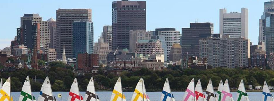 Colorful sailboats on the water with a backdrop of tall city skyscrapers and apartment buildings under a clear blue sky.