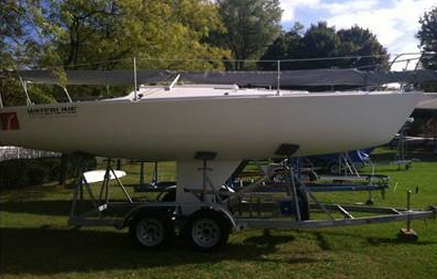 A white sailboat is mounted on a metal trailer parked on grass, with trees and other boats in the background under a partly sunny sky.