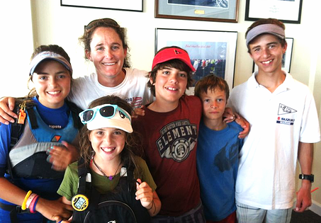 A group of six people, including four boys and two girls, smile together indoors. Some wear visors and casual clothing. They stand closely with arms around each other, with framed photos hanging on the wall behind them.