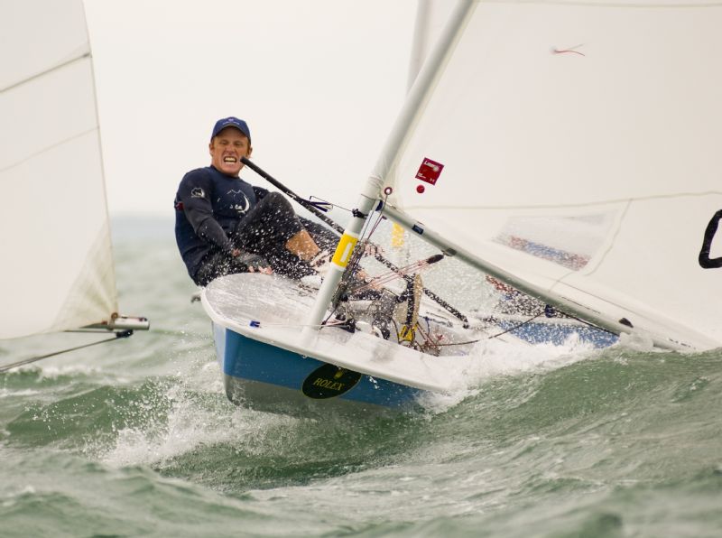 A person sailing a small dinghy on choppy water, leaning back and gripping the controls, with water spraying around the boat. The sailor is wearing a blue cap and wetsuit, and appears focused and determined.