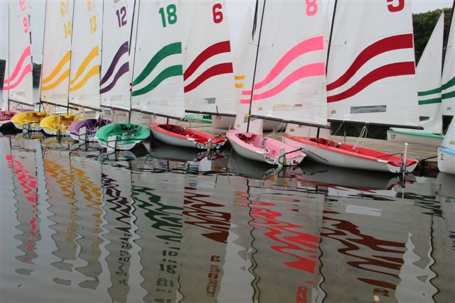 A row of colorful sailboats with striped sails is docked at the edge of calm water, their reflections creating wavy, vibrant patterns on the surface. The sails are marked with numbers and bold, colored stripes.