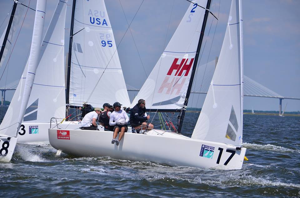 Four people sail a white racing sailboat numbered 17 in choppy water. Other sailboats and a large bridge appear in the background under a partly cloudy sky. The crew is actively maneuvering the boat.