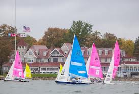 Several small sailboats with colorful blue, yellow, and pink sails race near the shore, with large houses and autumn trees in the background under a cloudy sky. An American flag is visible near the houses.