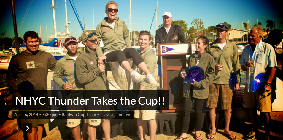 A group of sailors celebrates, some carrying one person. They are at a harbor with sailboats.