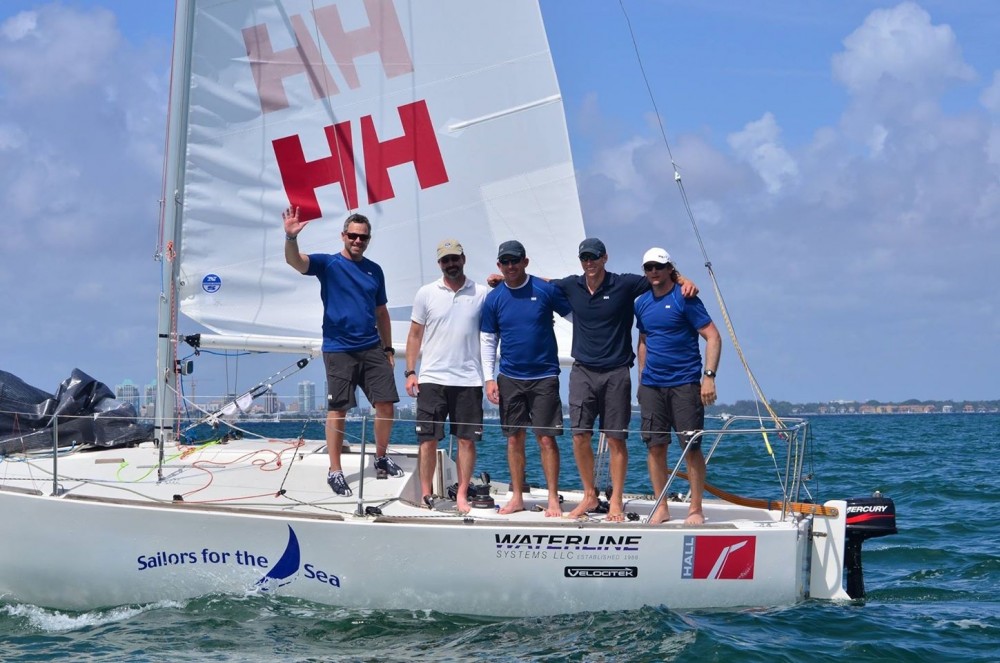 Five people stand on a white sailboat in the water, smiling at the camera. Four wear matching blue shirts and shorts, one wears a white shirt. The boat’s sail displays 'HH' in red, and city buildings are visible in the background.