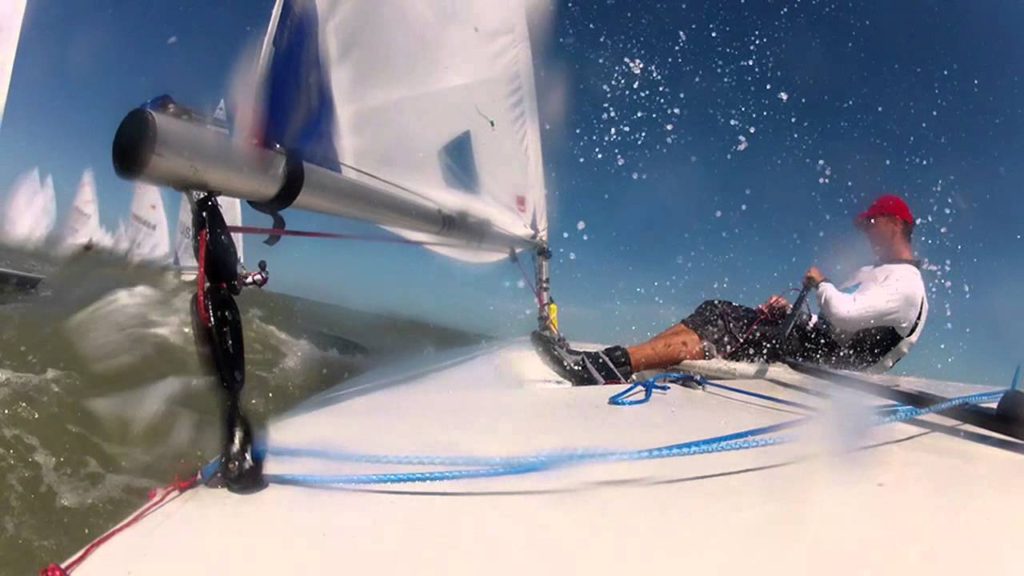 Two people sail a small boat on the water. One person leans back, holding a rope, while the other stands near the mast. Water splashes onto the deck and the sky is clear and blue.