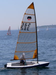 A person sails a small, single-mast sailboat with a clear sail marked '362 SAE' on a calm body of water, with other sailboats and distant shoreline visible in the background.