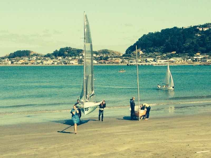 People stand by small sailboats on a sandy beach with calm blue water and distant hills. Some boats are in the water, while town buildings are visible across the bay under a partly cloudy sky.