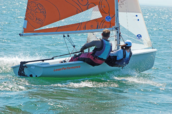Two people in life jackets sail a small boat labeled 'LaserPerformance' across clear blue water on a sunny day. The boat has an orange and white sail and creates small waves as it moves.